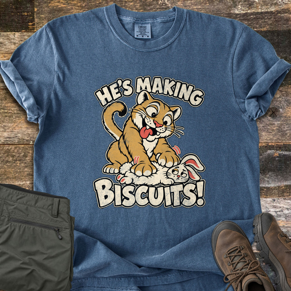 He's Making Biscuits Comfort Colors T-shirt in Denim, laid flat. A cartoon cougar with its tongue out kneads a dazed white rabbit with its paws, between the words He's Making Biscuits! It lies on weathered wood beside olive cargo pants and brown leather hiking boots.