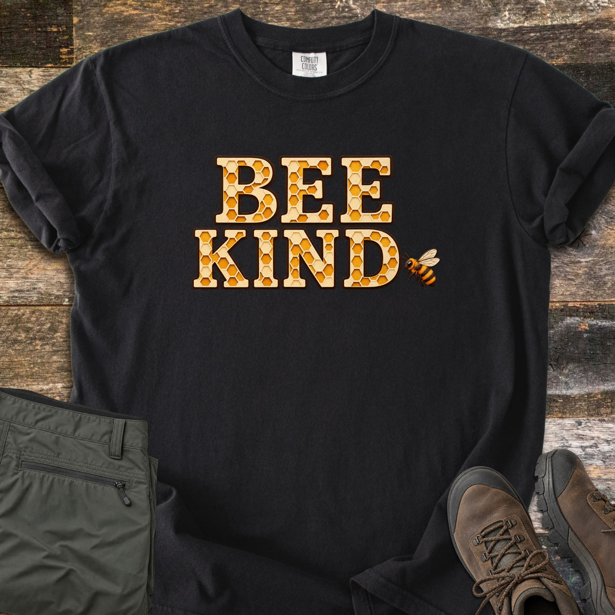 Bee Kind Comfort Colors T-shirt in Black, laid flat. The words BEE KIND in large letters filled with a golden honeycomb pattern, with a small bee beside them. Sleeves are rolled, on a weathered wood floor beside folded olive cargo pants and brown hiking boots.