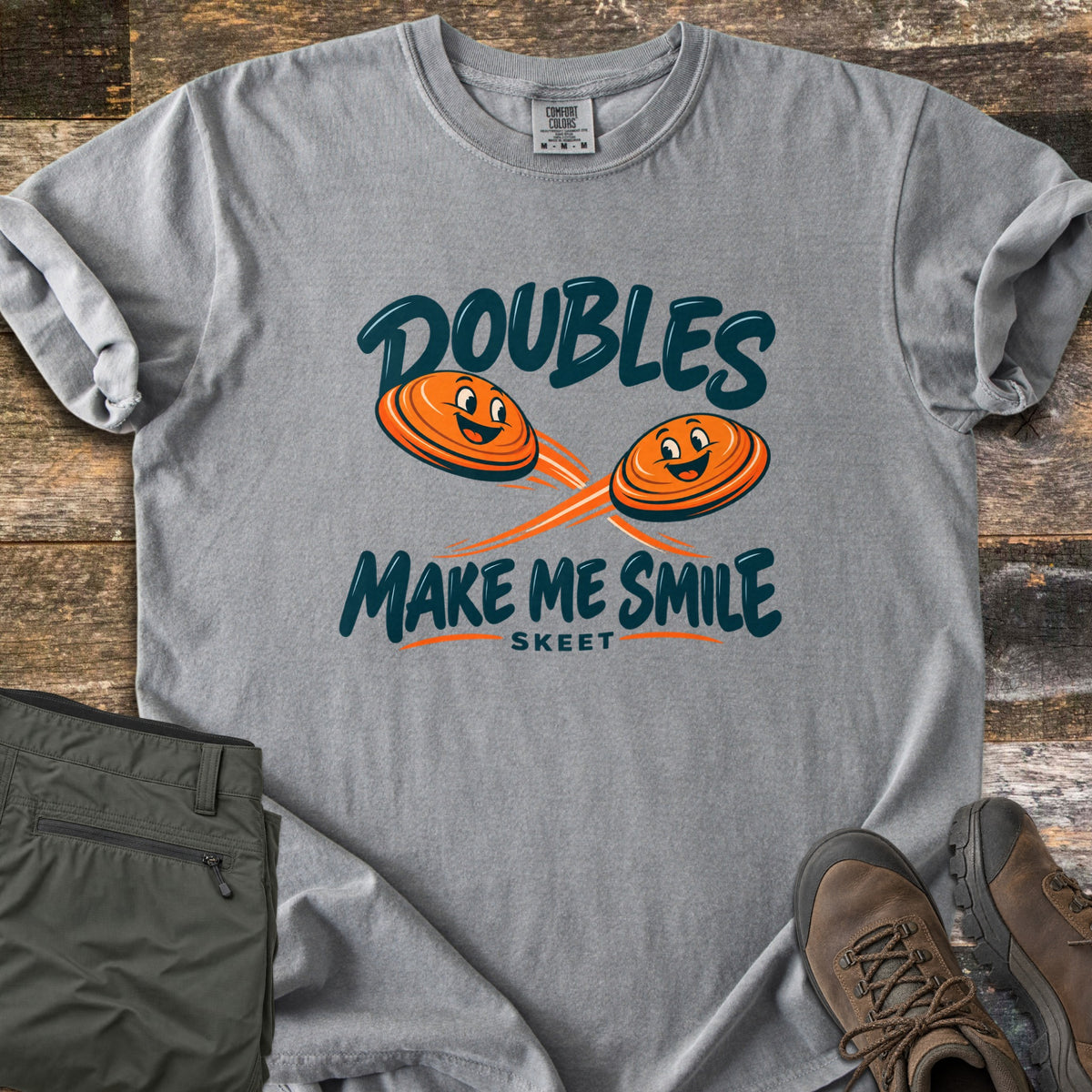 Doubles Make Me Smile Comfort Colors T-shirt in Grey, laid flat. Two smiling orange clay targets fly on crossing paths between the words Doubles Make Me Smile, with Skeet printed below. Folded olive pants and brown hiking boots sit beside it on weathered wood planks.