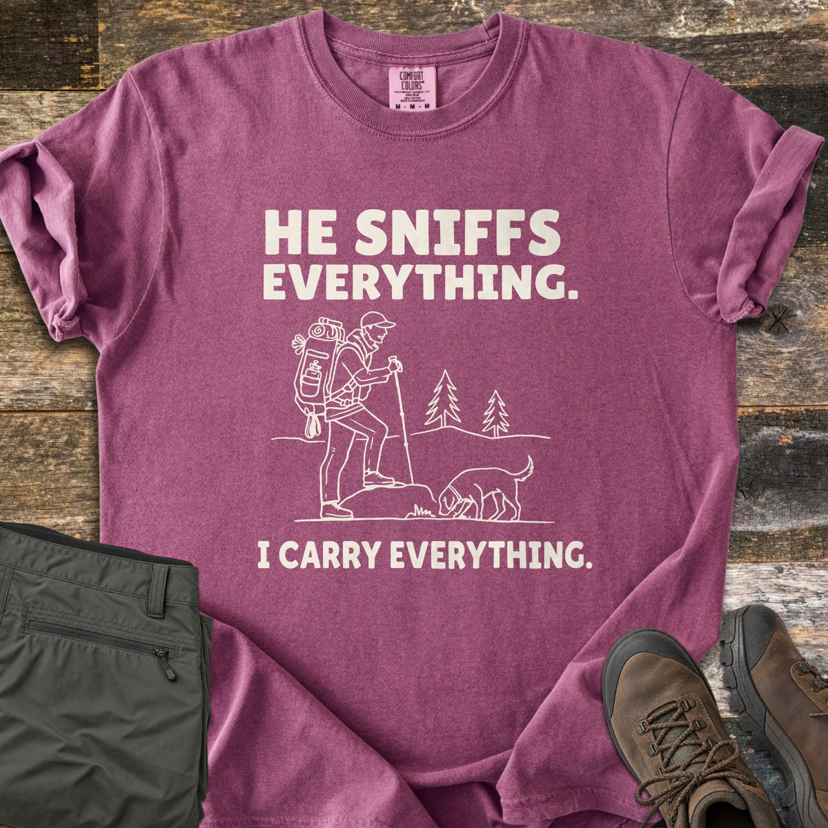 He Sniffs Everything. I Carry Everything Comfort Colors T-shirt in Berry, laid flat. Cream line art of a hiker with a large backpack and trekking pole beside a dog sniffing a rock, between the words He Sniffs Everything. and I Carry Everything. Olive pants and brown hiking boots sit on weathered wood.