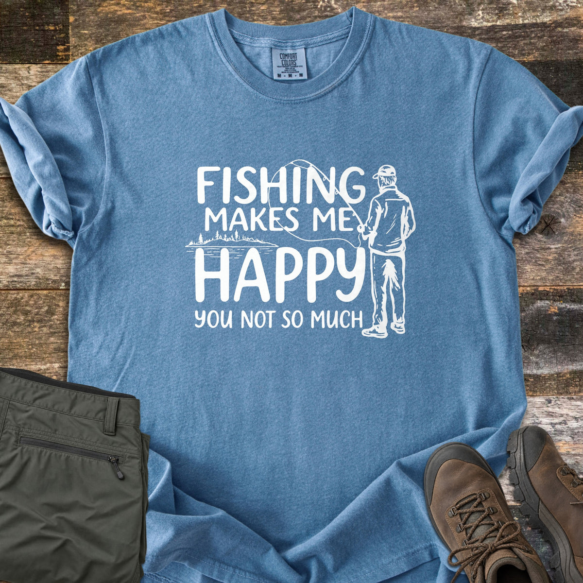 Fishing Makes Me Happy Comfort Colors T-shirt in Blue Jean, laid flat. White print of a man in a cap casting a fishing rod by a tree-lined lake, with the words Fishing Makes Me Happy You Not So Much. Laid on weathered wood with olive pants and brown hiking boots.