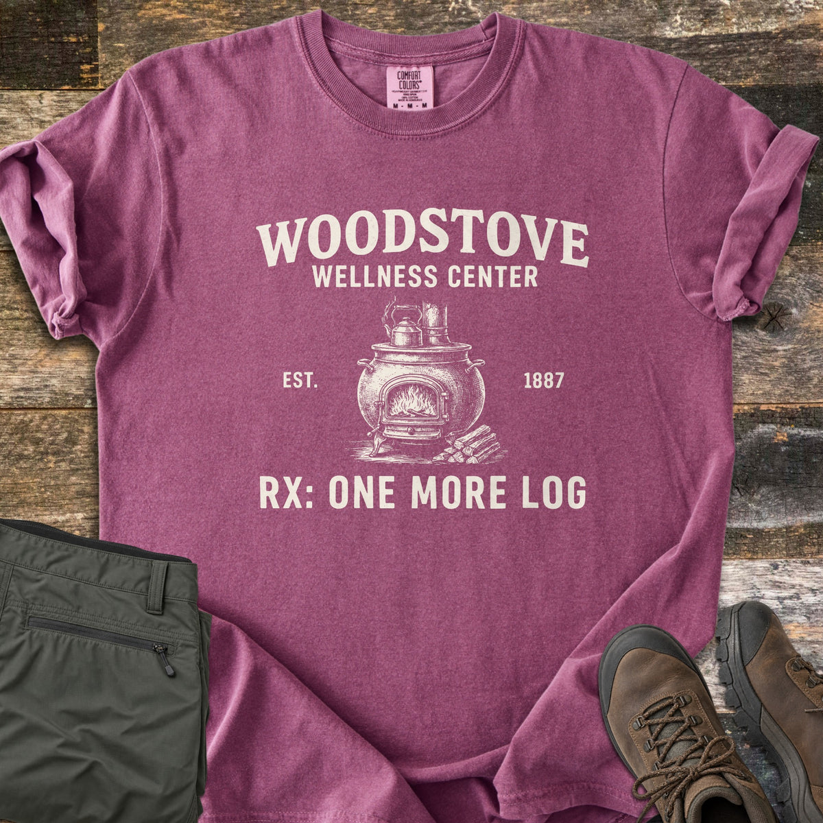 Woodstove Wellness Center Comfort Colors T-shirt in Berry, laid flat. Cream print of a potbelly woodstove with a fire, a kettle on top and logs beside it, under Woodstove Wellness Center, flanked by Est. 1887, above Rx: One More Log. On wood planks with olive pants and brown hiking boots.