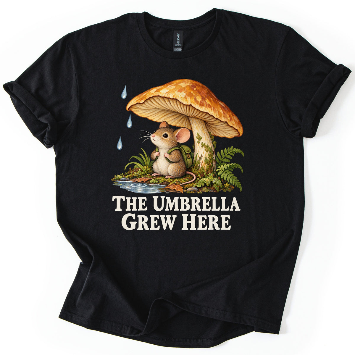 The Umbrella Grew Here Comfort Colors T-shirt in Black, laid flat. A mouse wearing a green backpack sits under a large orange mushroom as raindrops fall, beside ferns and a puddle, above the words The Umbrella Grew Here in cream letters. Shown on a white background.