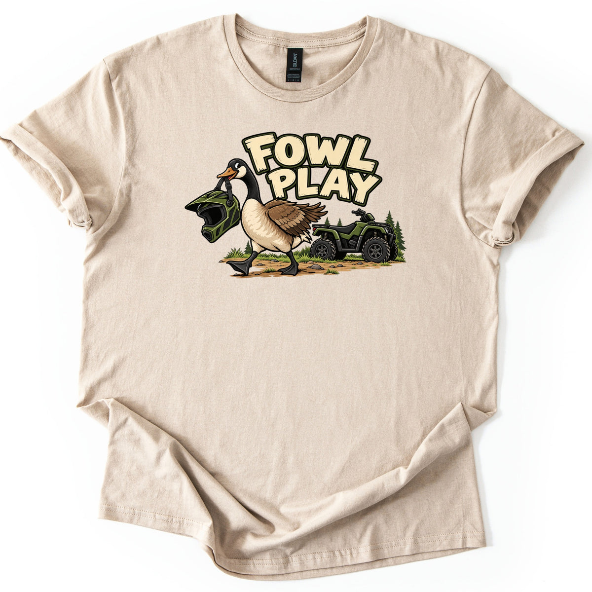 Fowl Play Comfort Colors T-shirt in Sand, laid flat. A cartoon Canada goose carries a green motocross helmet in its beak past a green ATV and pine trees, below the words Fowl Play. The shirt lies on a white background with cuffed sleeves.