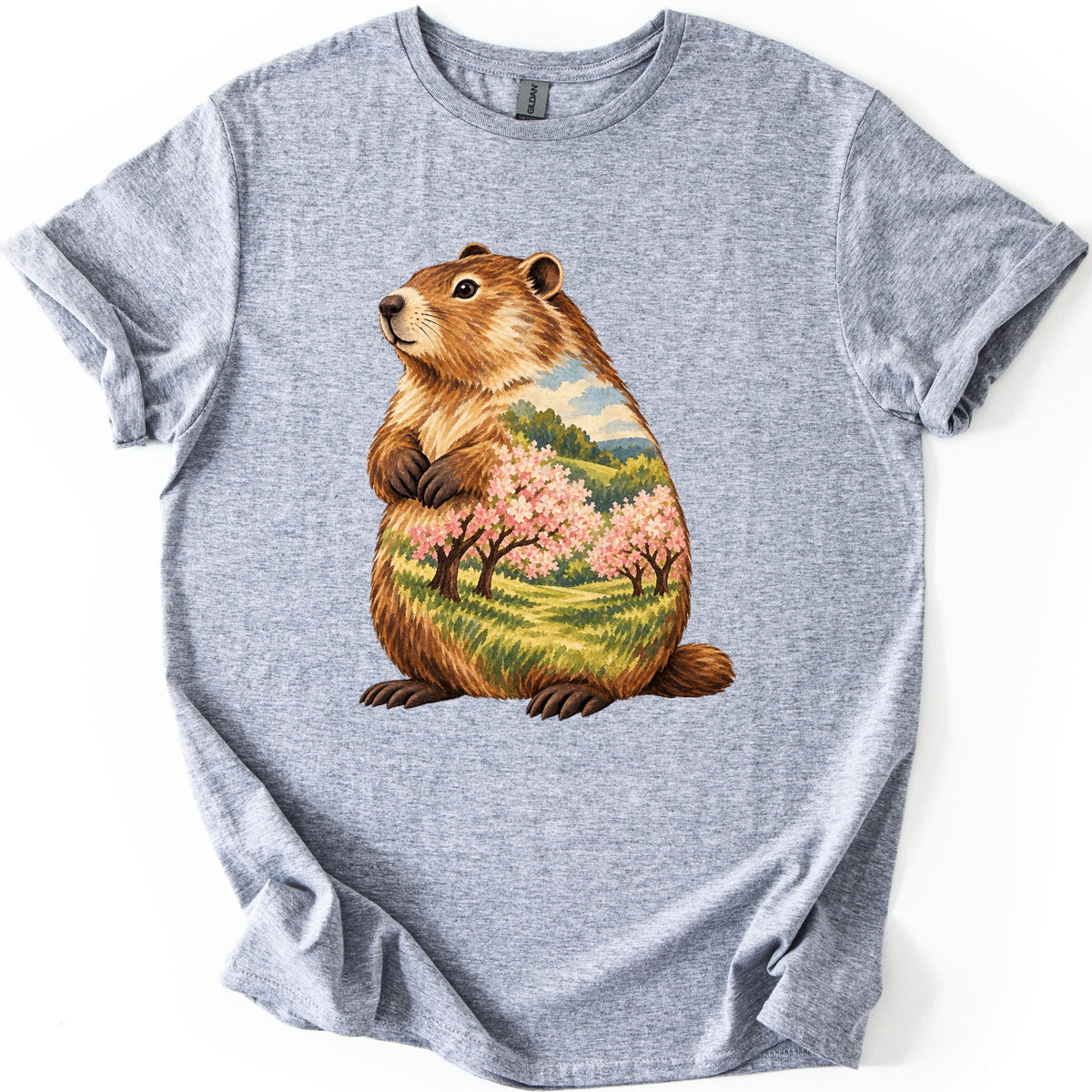 Groundhog Blossom Meadow Comfort Colors T-shirt in Sport Grey, laid flat. A painted groundhog sits upright with paws at its chest, its body filled with a meadow of pink blossoming trees, green hills and blue sky. No words. Sleeves cuffed, on a white background.