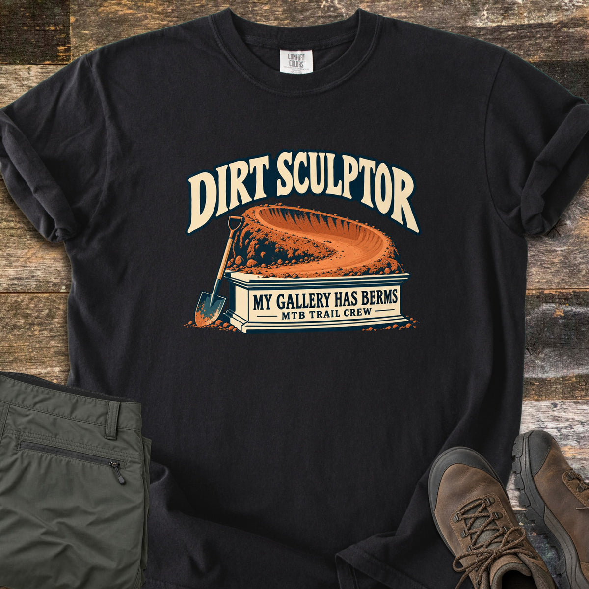 Dirt Sculptor Comfort Colors T-shirt in Black, laid flat. The words Dirt Sculptor arch over a dirt berm on a stone pedestal reading My Gallery Has Berms, MTB Trail Crew, with a shovel leaning on it. Olive pants and brown hiking boots lie on wood planks.