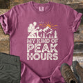 My Kind Of Peak Hours Comfort Colors T-shirt in Berry, laid flat. A cream hiker with a backpack sits on a rock with a steaming mug, facing mountains, pines and an orange sun, above the words My Kind Of Peak Hours. Olive hiking pants and brown boots lie on weathered wood.