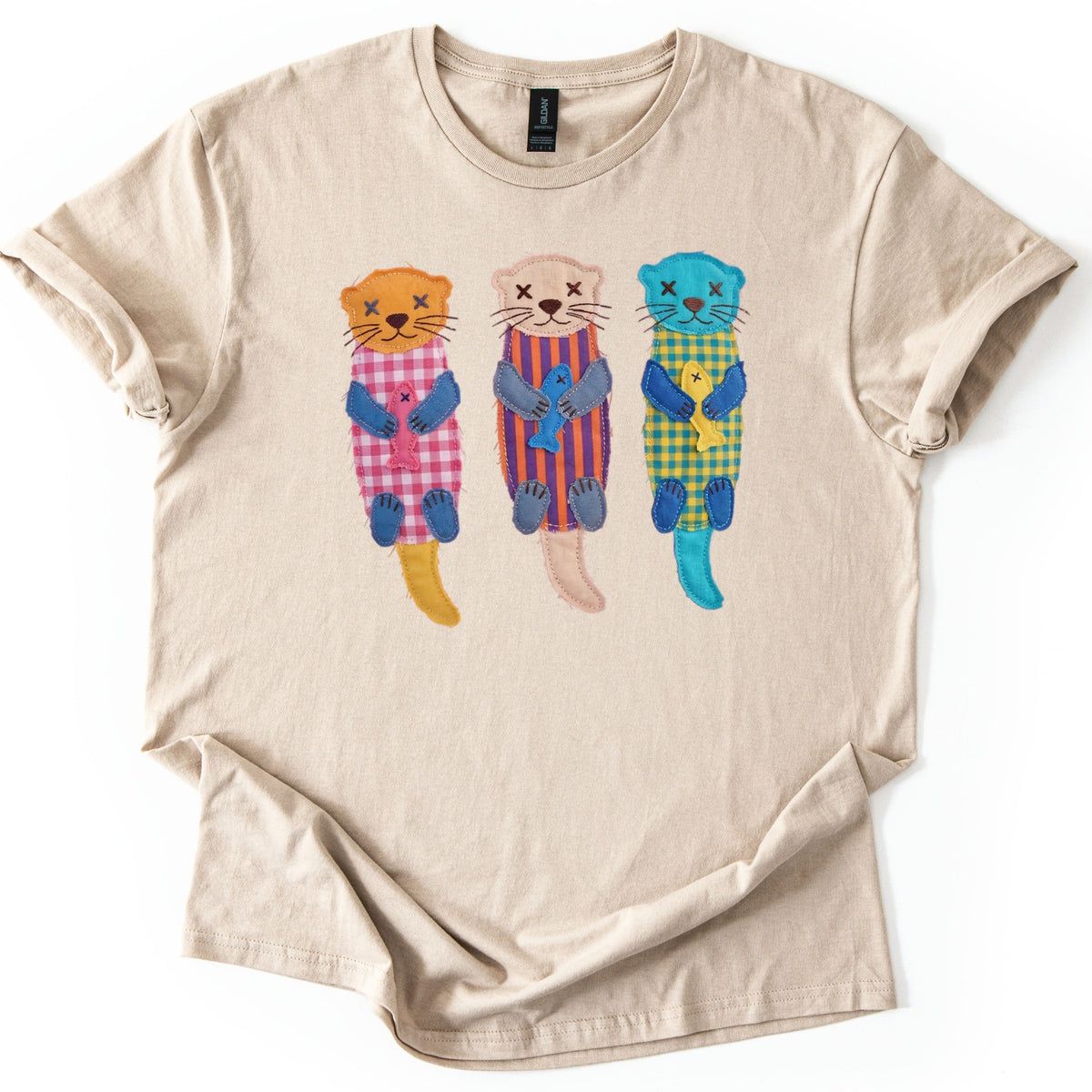 Patchwork Otter Comfort Colors T-shirt in Sand, laid flat. Three stitched patchwork otters float on their backs holding fish: orange with pink gingham, beige with purple and orange stripes, turquoise with yellow gingham. No words. Cuffed sleeves on a white surface.