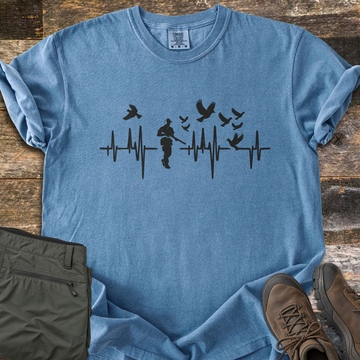 Bird Hunter Comfort Colors T-shirt in Blue Jean, laid flat. A black heartbeat line crosses the chest with a hunter silhouette in a cap holding a shotgun and birds flying up from the line. Folded olive pants and brown hiking boots lie beside it on weathered wood.