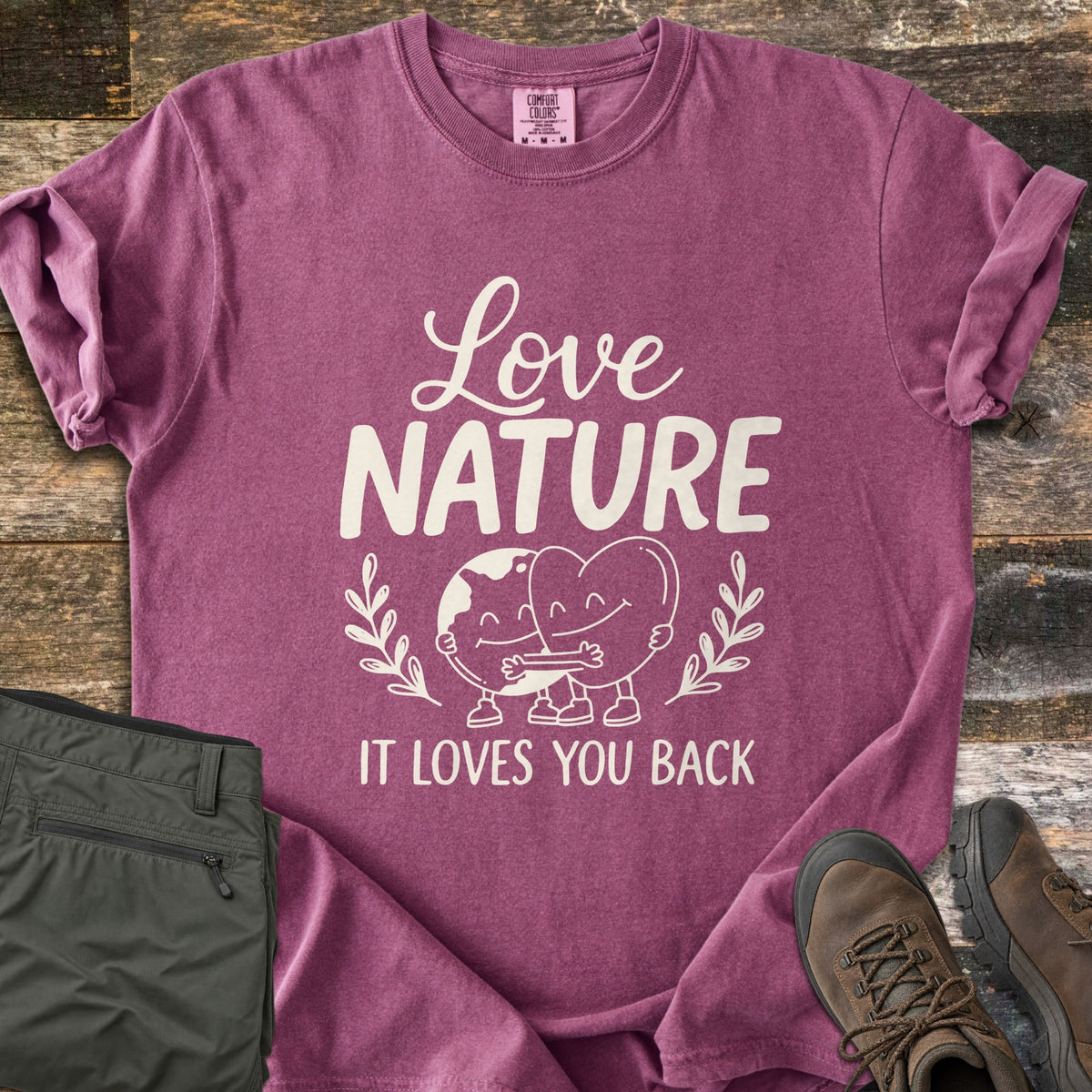 Nature Loves You Back Comfort Colors T-shirt in Berry, laid flat. Cream print reads Love Nature above a smiling cartoon Earth and heart hugging between leafy sprigs, with It Loves You Back below. It lies on weathered wood beside olive hiking pants and brown boots.
