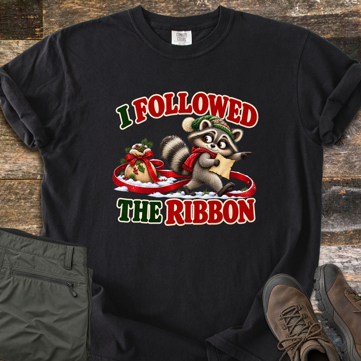 I Followed The Ribbon Comfort Colors T-shirt in Black, laid flat. A cartoon raccoon in a green knit hat and red scarf holds a map and points ahead, walking along a red ribbon from a gift sack in snow, between the words I Followed The Ribbon. Olive pants and brown boots lie on a wood floor.
