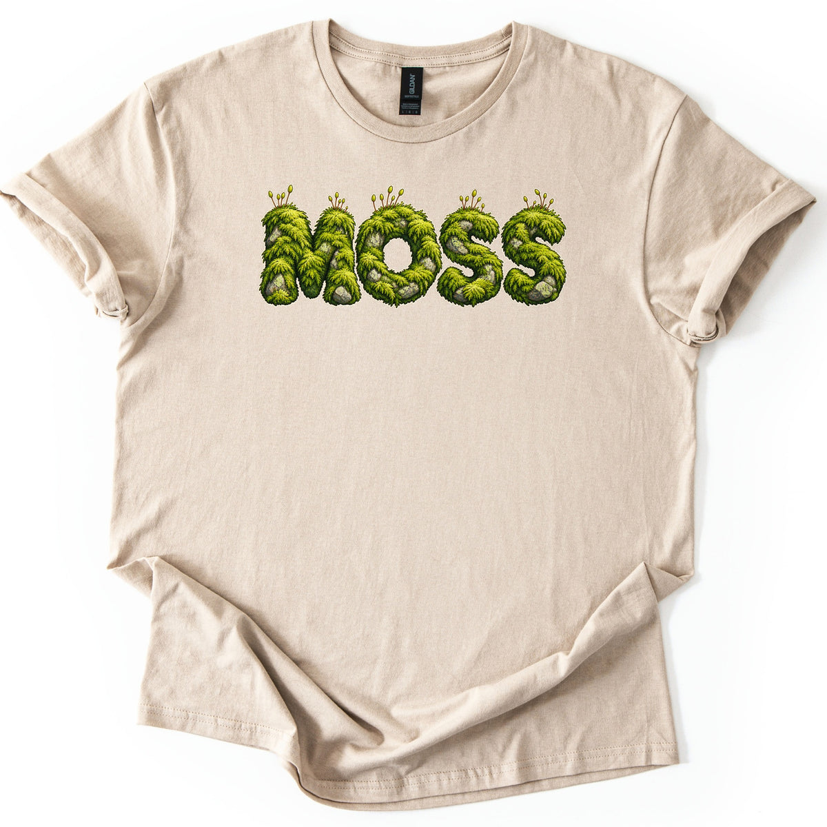 Moss Comfort Colors T-shirt in Sand, laid flat. The word MOSS is printed in fuzzy green moss-covered letters with gray stones and tiny sprouting stalks along the top. The shirt has rolled sleeves and a folded hem on a white background.