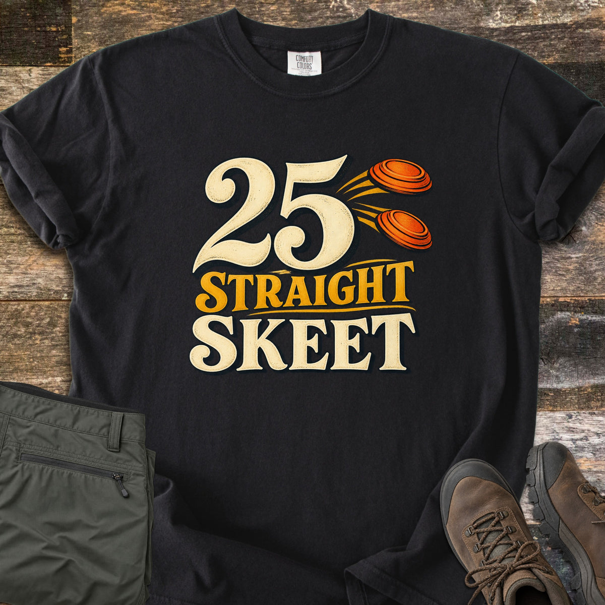25 Straight Skeet Comfort Colors T-shirt in Black, laid flat. Large cream numbers 25 with two orange clay targets flying beside them, above the words Straight Skeet in gold and cream. Sleeves are rolled on a wood plank surface with olive cargo pants and brown hiking boots.