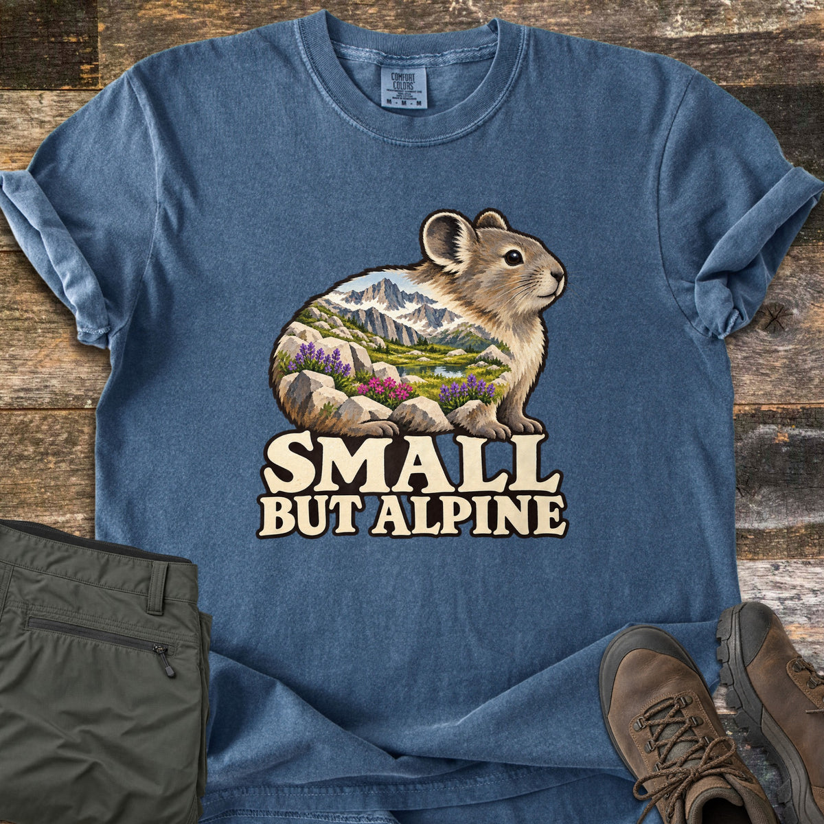 Small But Alpine Comfort Colors T-shirt in Denim, laid flat. A pika on rocks, its body filled with a scene of snowy peaks, a lake and purple and pink wildflowers, above the words Small But Alpine. Olive hiking pants and brown hiking boots sit on a wood floor.