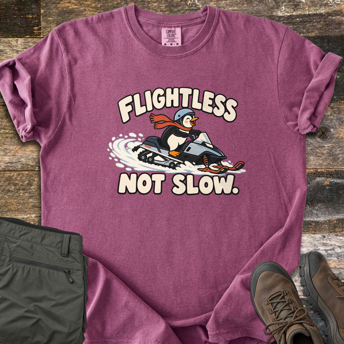 Flightless Not Slow Comfort Colors T-shirt in Berry, laid flat. A cartoon penguin in a helmet and red scarf rides a snowmobile through snow between the words Flightless Not Slow. Folded olive pants and brown hiking boots sit beside it on weathered wood.
