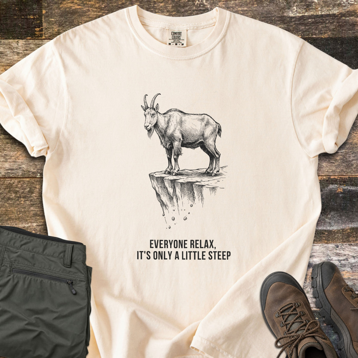 Everyone Relax It's Only A Little Steep Comfort Colors T-shirt in Ivory, laid flat. A black sketch of a horned mountain goat standing on a crumbling cliff ledge above the words Everyone Relax, It's Only A Little Steep. Sleeves are rolled, on wood planks with green hiking pants and brown boots.