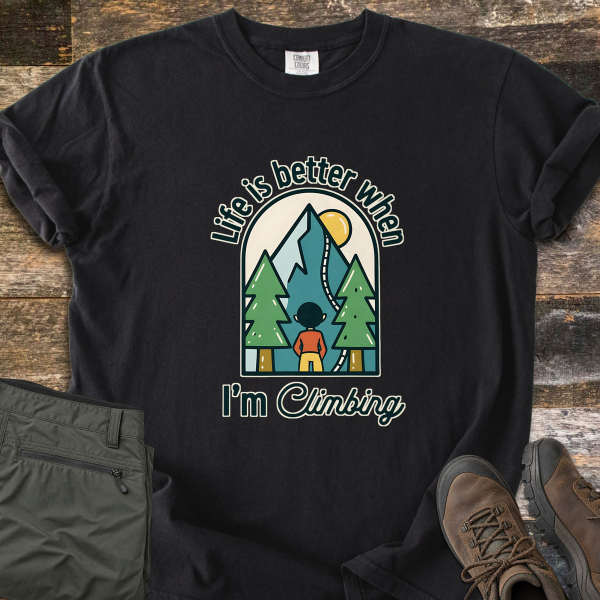 Better When I'm Climbing Comfort Colors T-shirt in Black, laid flat. An arched scene of a person facing a mountain with a winding trail, pine trees and a yellow sun, with the words Life is better when I'm Climbing. Gray pants and brown hiking boots lie on a wood floor.