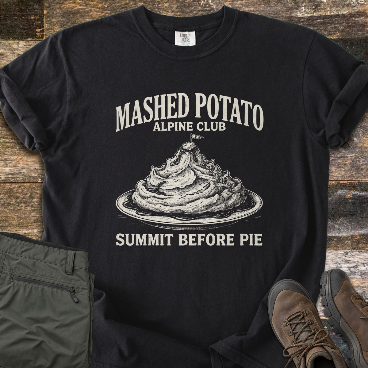 Mashed Potato Alpine Club Comfort Colors T-shirt in Black, laid flat. An etched drawing of a mashed potato mountain on a plate, with tiny climbers and a flag on a butter pat summit, under Mashed Potato Alpine Club and above Summit Before Pie. Green hiking pants and boots sit on wood.