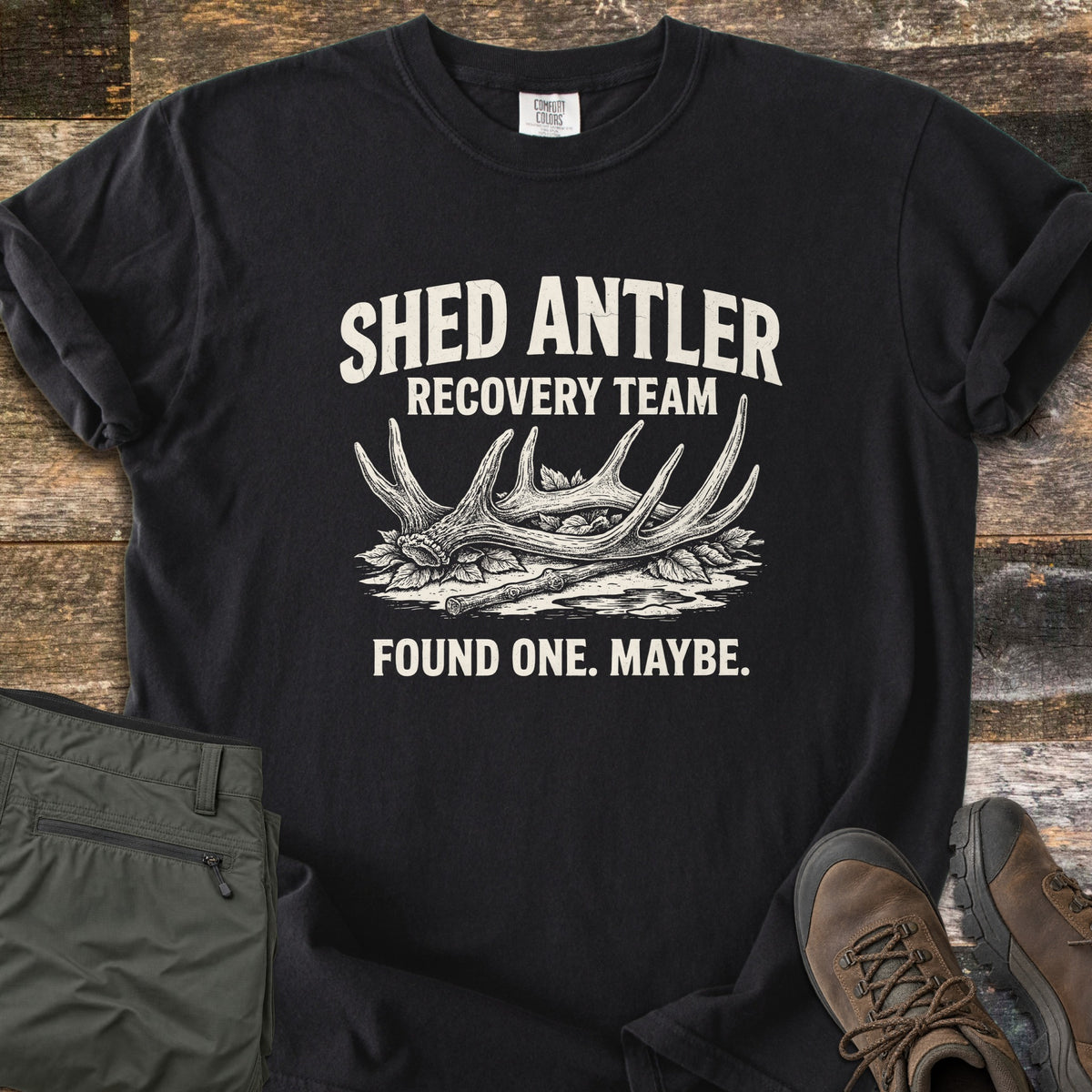 Shed Antler Recovery Team Comfort Colors T-shirt in Black, laid flat. A sketched shed antler rests on leaves beside a stick under the words Shed Antler Recovery Team, with Found One. Maybe. below. Olive pants and brown hiking boots sit beside it on weathered wood.