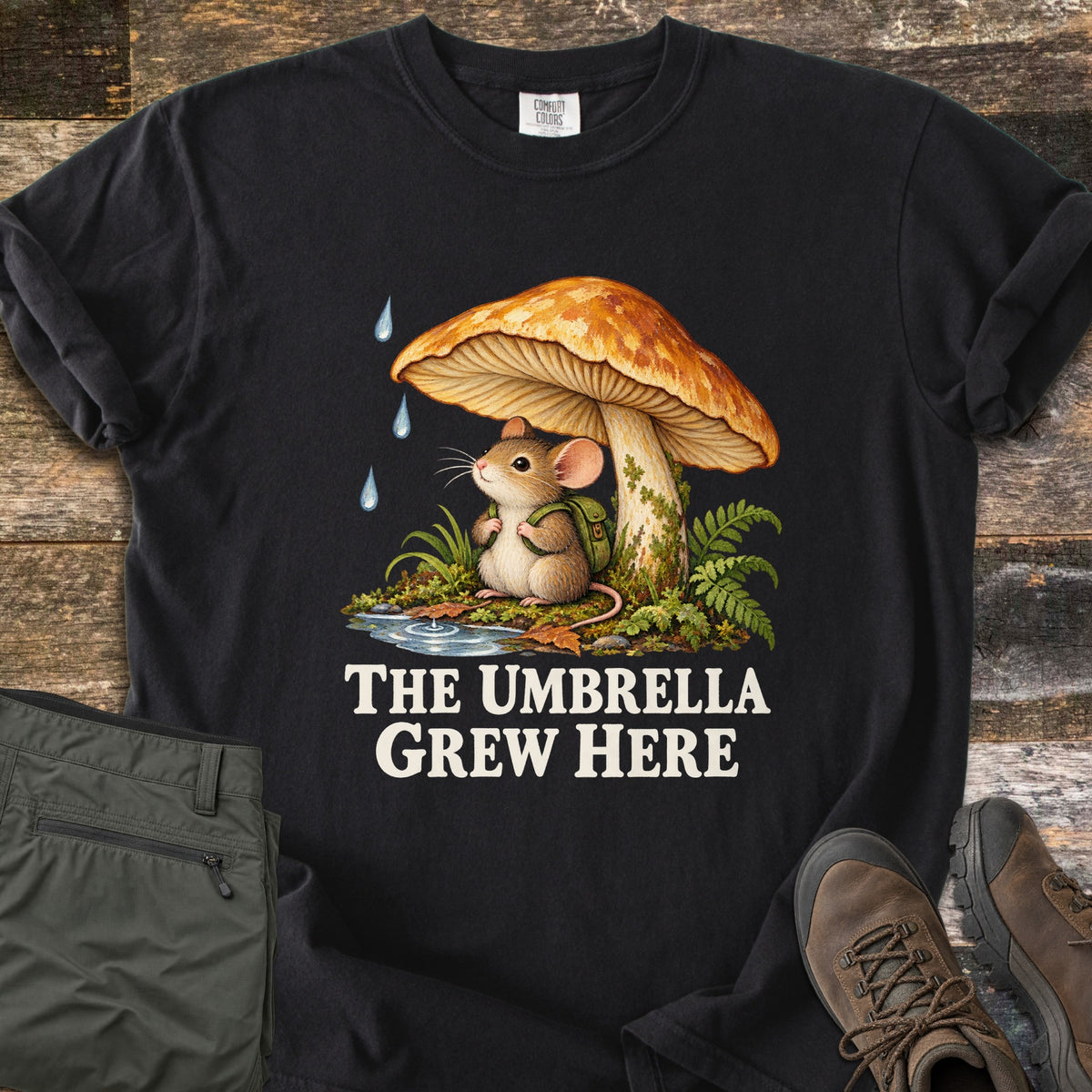 The Umbrella Grew Here Comfort Colors T-shirt in Black, laid flat. A mouse wearing a green backpack shelters under a large orange mushroom from falling raindrops, above the words The Umbrella Grew Here in cream lettering. Olive pants and brown hiking boots lie beside it on weathered wood.