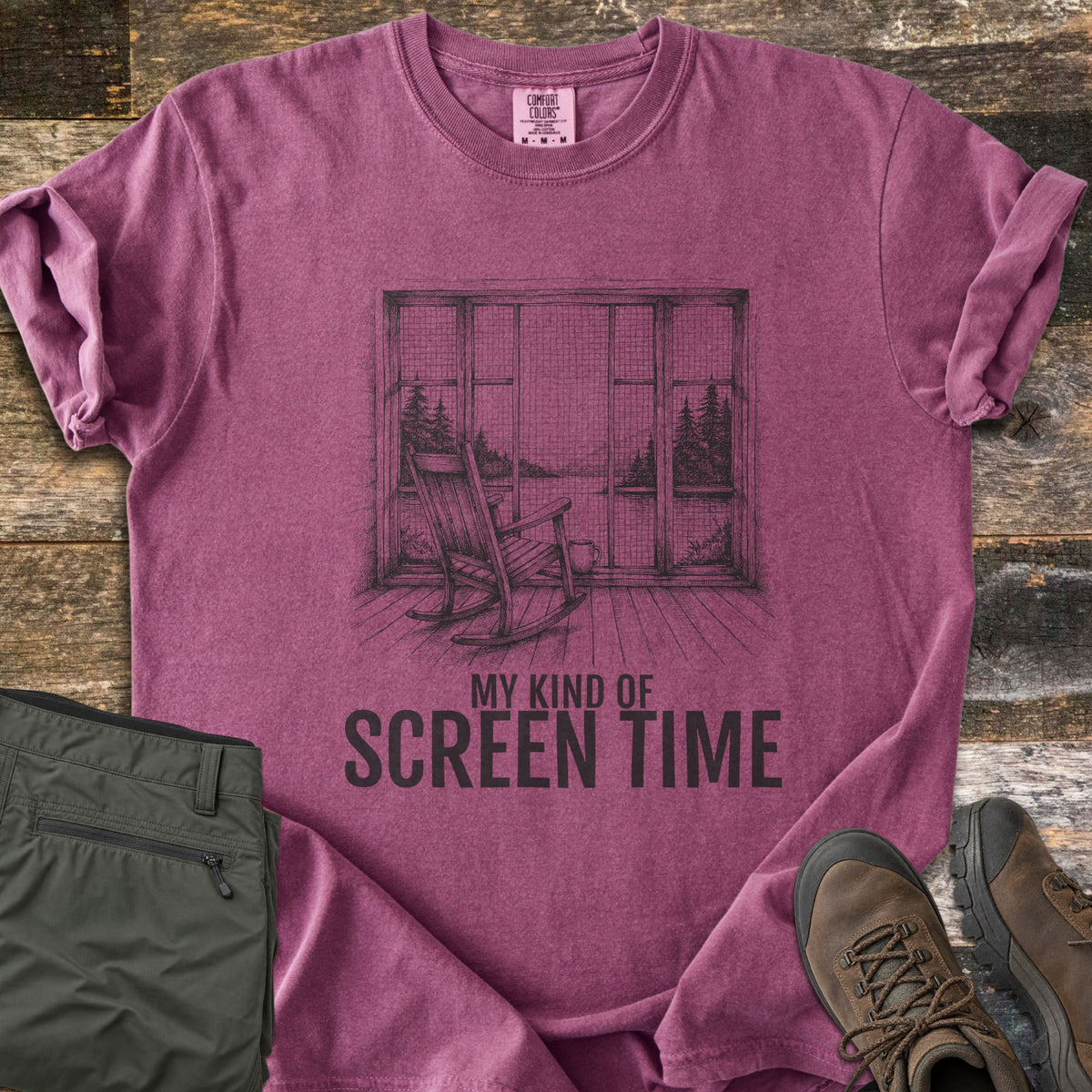My Kind Of Screen Time Comfort Colors T-shirt in Berry, laid flat. A sketched rocking chair on a plank porch faces a screened window overlooking a lake framed by pines, with a mug beside it, above the words My Kind Of Screen Time. Olive hiking pants and brown boots lie on weathered boards.