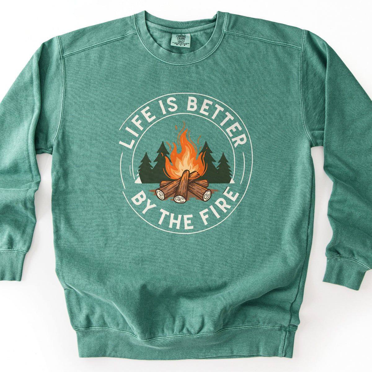 Life Is Better By The Fire Ring Comfort Colors crewneck sweatshirt in Light Green, laid flat. A campfire of stacked logs with orange flames sits in front of dark green pines inside a cream ring, with the words Life Is Better By The Fire curved around it. It lies on a white surface.