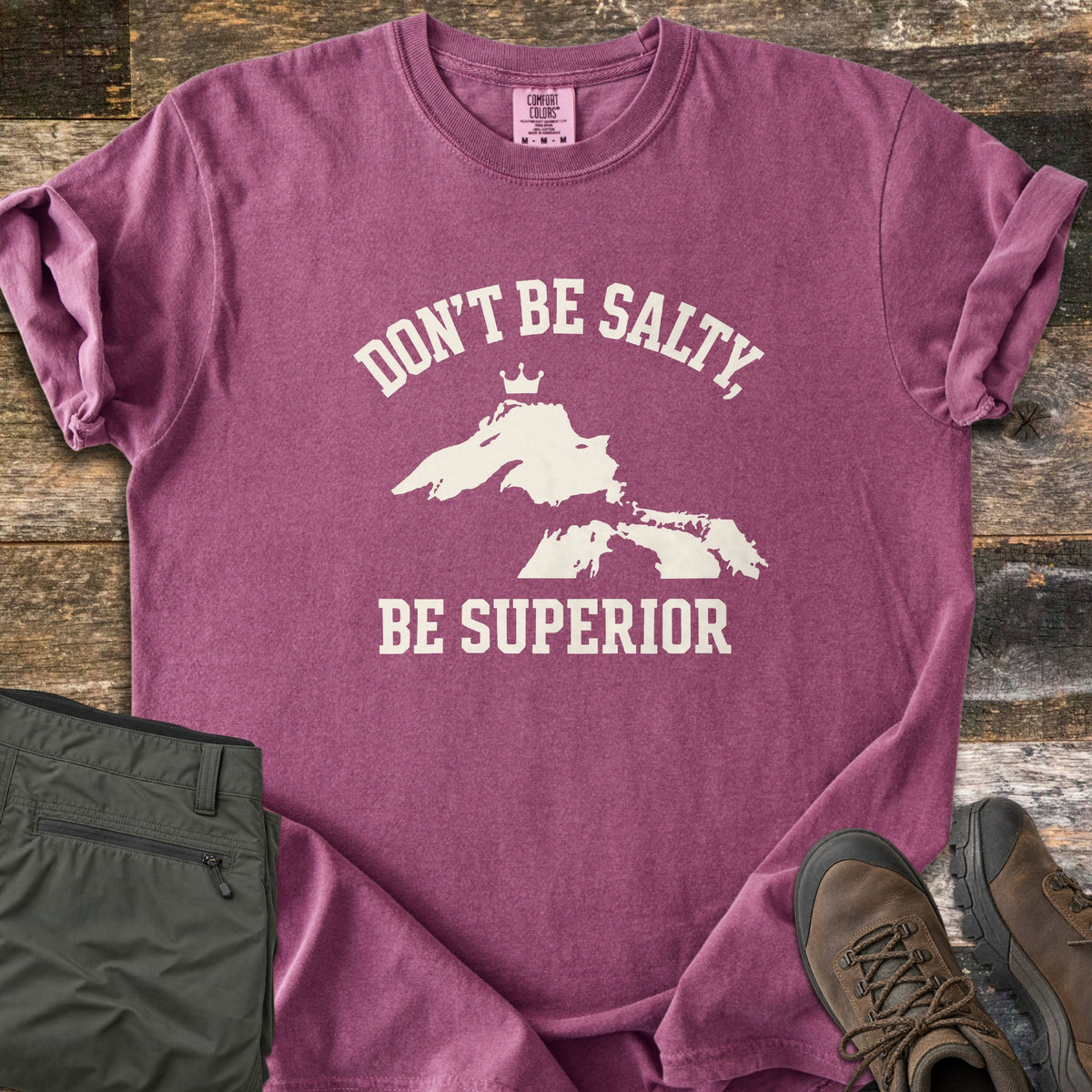 Be Superior Comfort Colors T-shirt in Berry, laid flat. Cream text reads Don't Be Salty, arched over a Lake Superior silhouette topped with a small crown, with Be Superior below. Sleeves rolled, on weathered wood with olive hiking pants and brown hiking boots.
