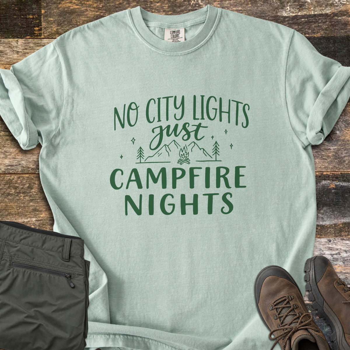 Campfire Nights Comfort Colors T-shirt in Bay, laid flat. Dark green text reads No City Lights just Campfire Nights around line art of mountains, pine trees, stars and a campfire. Sleeves rolled, on weathered wood with olive hiking pants and brown boots.