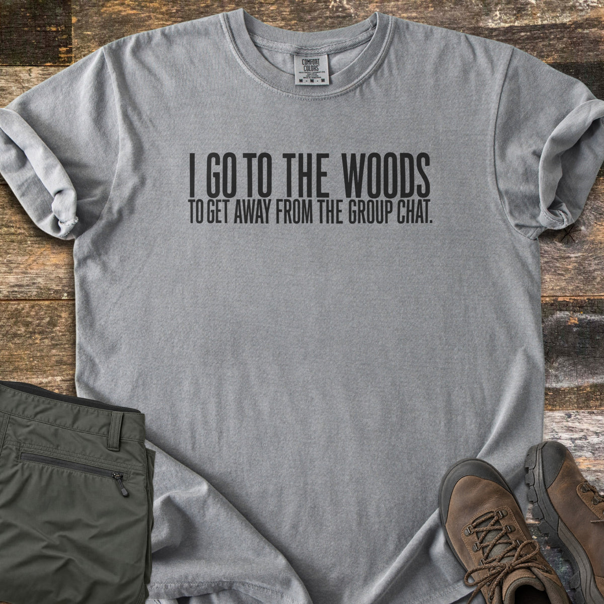 Away From The Group Chat Comfort Colors T-shirt in Grey, laid flat. Black block letters read I GO TO THE WOODS above TO GET AWAY FROM THE GROUP CHAT. The shirt has rolled sleeves and lies on weathered wood boards beside folded olive green pants and brown hiking boots.