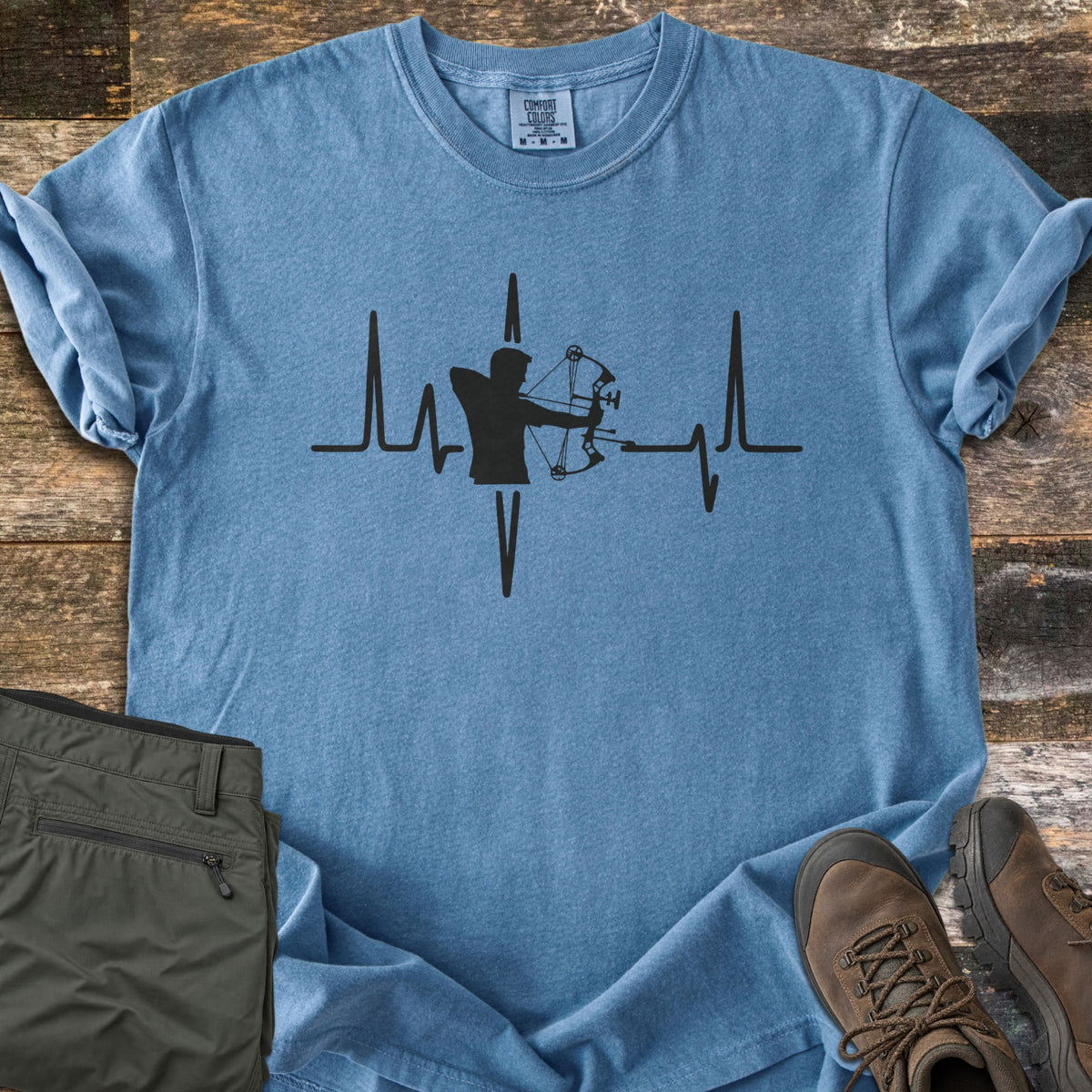 Compound Bow Heartbeat Comfort Colors T-shirt in Blue Jean, laid flat. A black heartbeat line with the silhouette of an archer drawing a compound bow at its center. Sleeves are rolled on a wood plank floor, with olive cargo pants and brown hiking boots beside it.