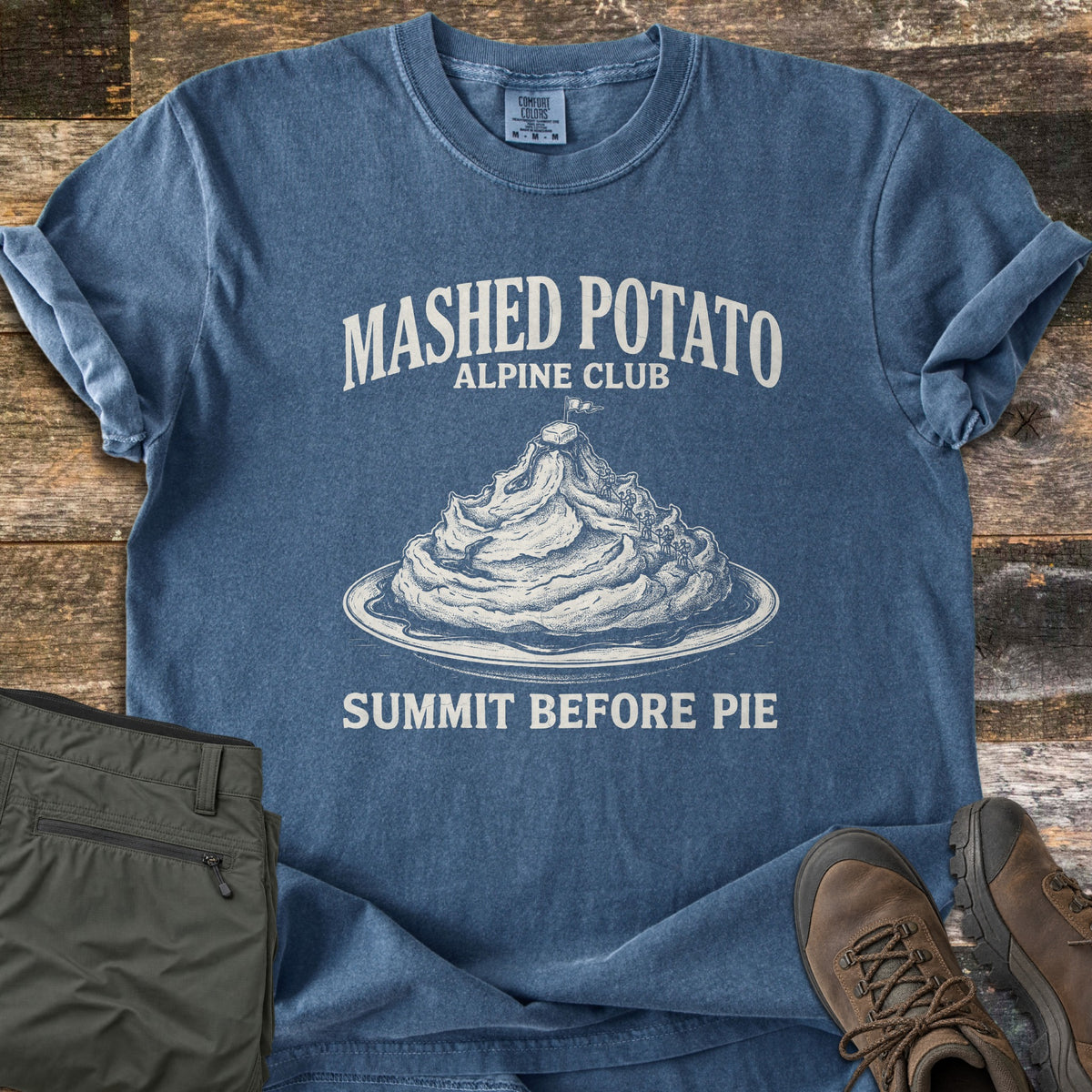 Mashed Potato Alpine Club Comfort Colors T-shirt in Denim, laid flat. A mound of mashed potatoes on a plate topped with a pat of butter and a flag, with tiny climbers ascending, under Mashed Potato Alpine Club and above Summit Before Pie. Olive hiking pants and brown boots sit on wood boards.