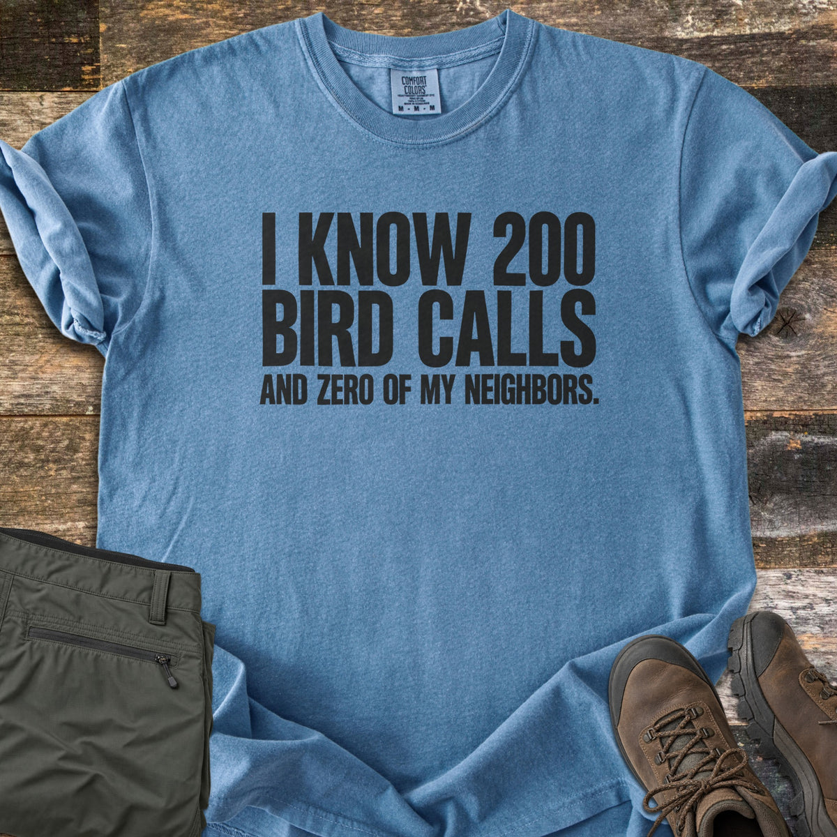 I Know 200 Bird Calls and Zero of My Neighbors Comfort Colors T-shirt in Blue Jean, laid flat. Bold black capital letters read I KNOW 200 BIRD CALLS AND ZERO OF MY NEIGHBORS. The shirt has rolled sleeves and lies on weathered wood boards beside folded olive hiking pants and brown hiking boots.