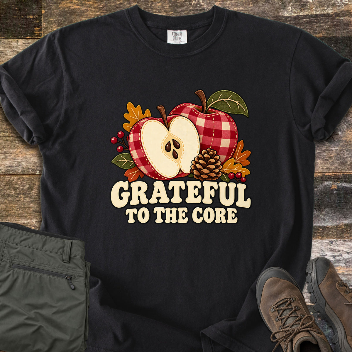 Grateful to the Core Comfort Colors T-shirt in Black, laid flat. A red gingham apple and an apple half with seeds sit with a pinecone, autumn leaves and berries above the words Grateful to the Core. Sleeves rolled, it lies on weathered wood by olive pants and brown boots.