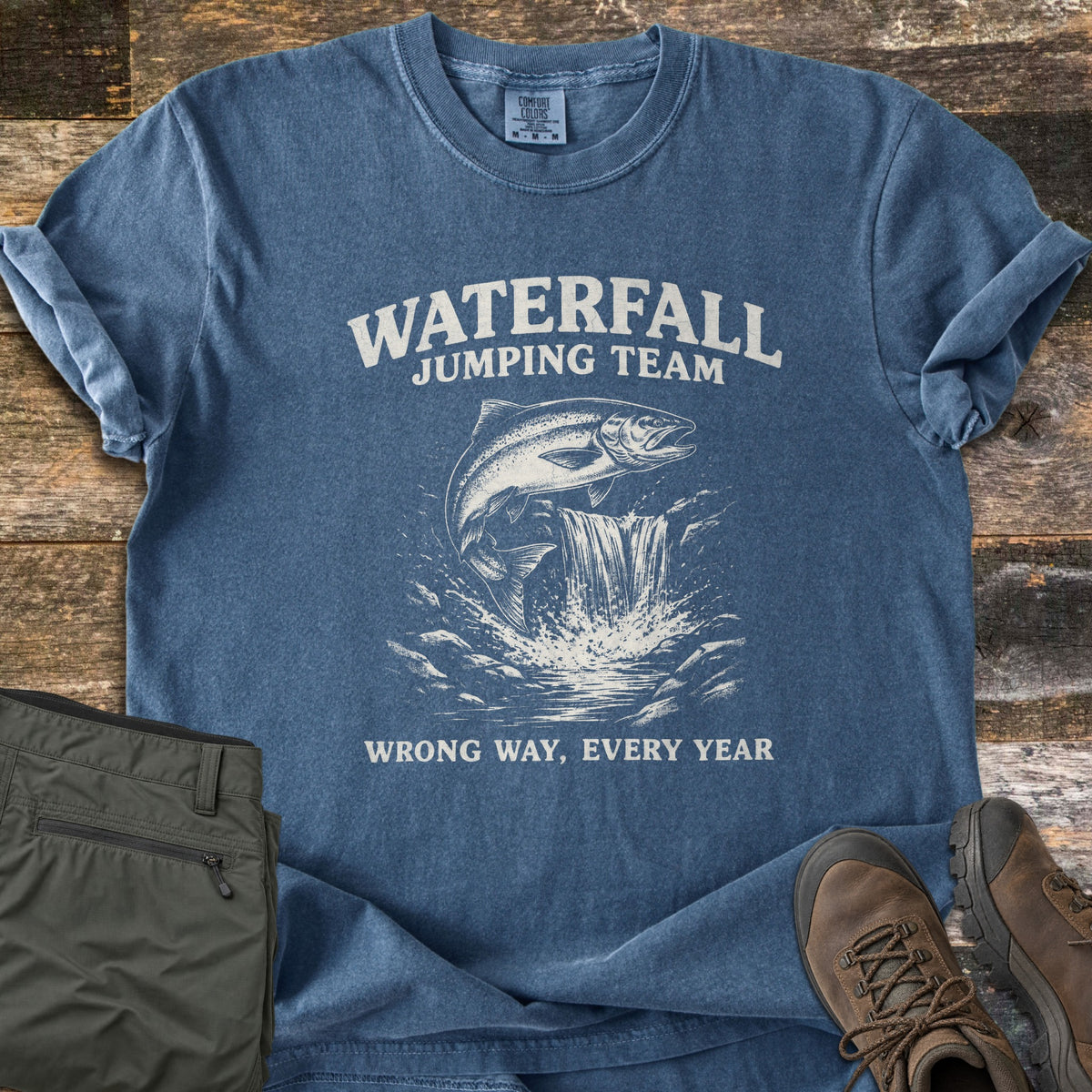 Waterfall Jumping Team Comfort Colors T-shirt in Denim, laid flat. A salmon leaps up a waterfall between the words Waterfall Jumping Team and Wrong Way, Every Year. Rolled sleeves, on weathered wood with olive pants and brown hiking boots.