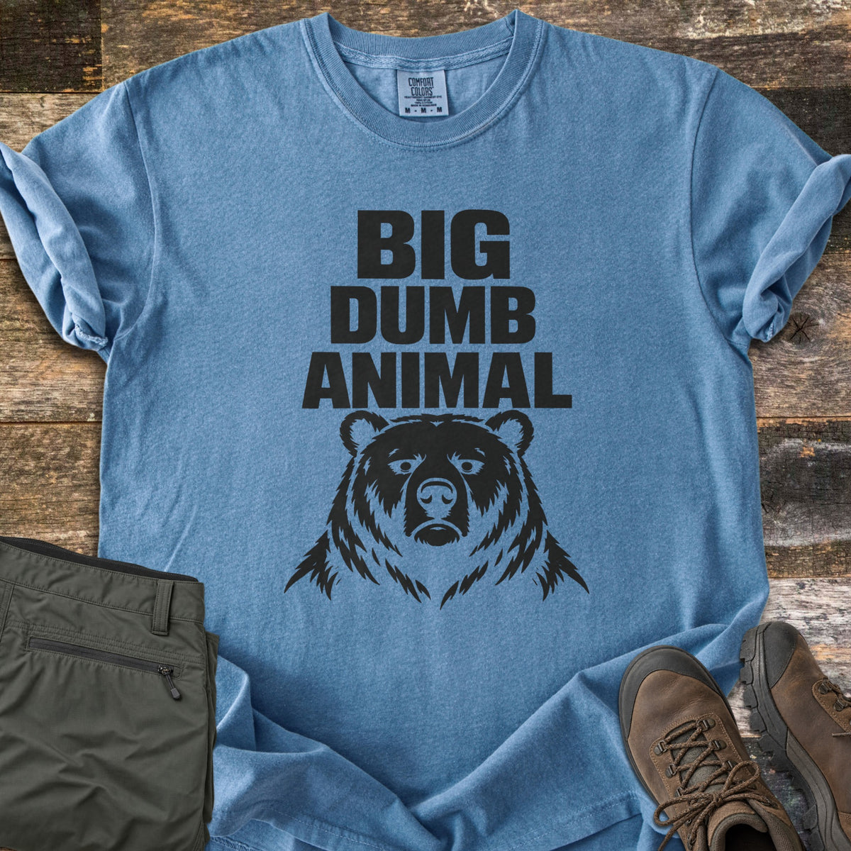Big Dumb Animal Comfort Colors T-shirt in Blue Jean, laid flat. Bold black block letters read Big Dumb Animal above a grumpy-faced bear head. The shirt, sleeves rolled, lies on weathered wood planks beside folded olive hiking pants and brown hiking boots.