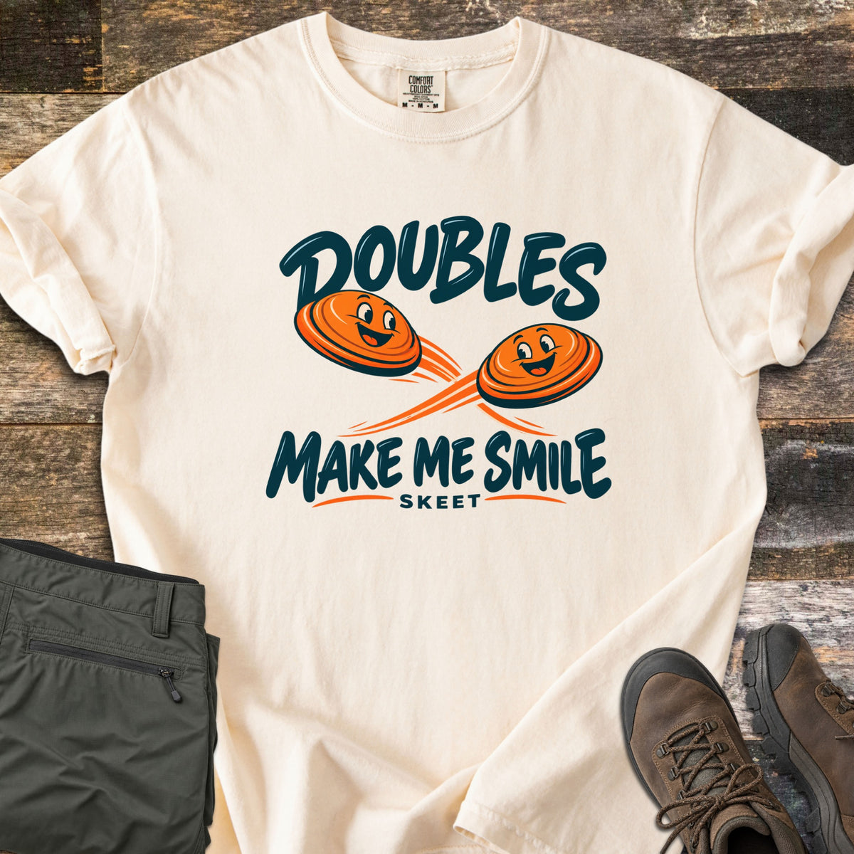Doubles Make Me Smile Comfort Colors T-shirt in Ivory, laid flat. Two smiling orange clay targets fly on crossing paths between the words Doubles Make Me Smile, with Skeet printed below. Folded olive pants and brown hiking boots sit beside it on weathered wood planks.