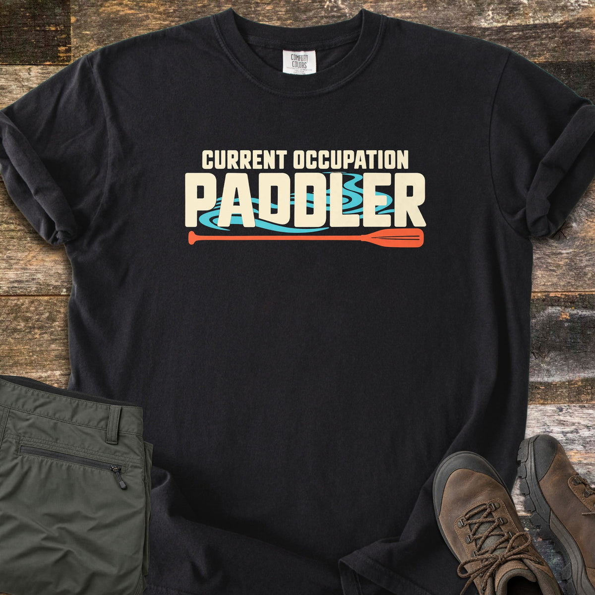 Current Occupation Paddler Comfort Colors T-shirt in Black, laid flat. Cream block letters read Current Occupation Paddler over turquoise water swirls, with an orange canoe paddle beneath. Sleeves are rolled on a weathered wood surface beside olive hiking pants and brown hiking boots.