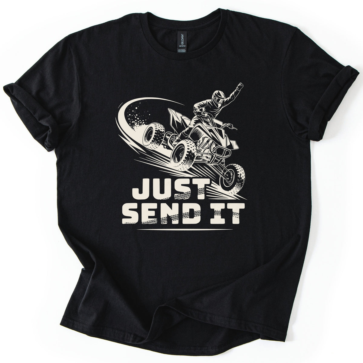 ATV Just Send It Comfort Colors T-shirt in Black, laid flat. A cream illustration of a helmeted rider on an airborne quad ATV, one fist raised, spraying dust above the words Just Send It in bold tire-tread letters. Sleeves are cuffed, on a white background.