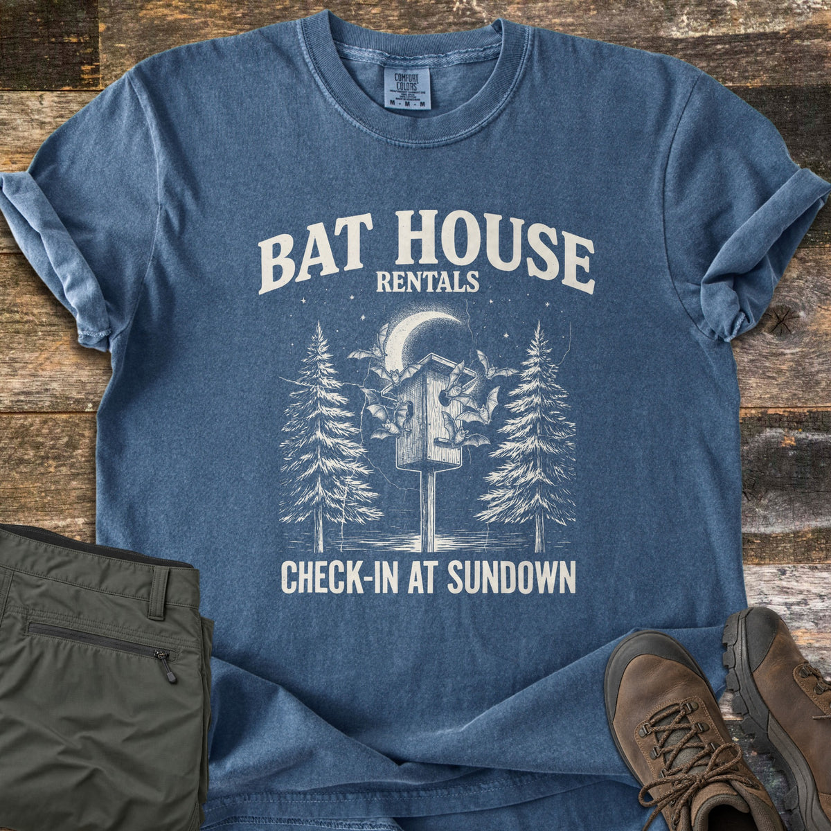 Bat House Rentals Comfort Colors T-shirt in Denim, laid flat. Words Bat House Rentals above a bat house on a post with bats, a crescent moon and pine trees, and Check-In At Sundown below. Olive pants and brown hiking boots sit on weathered wood.