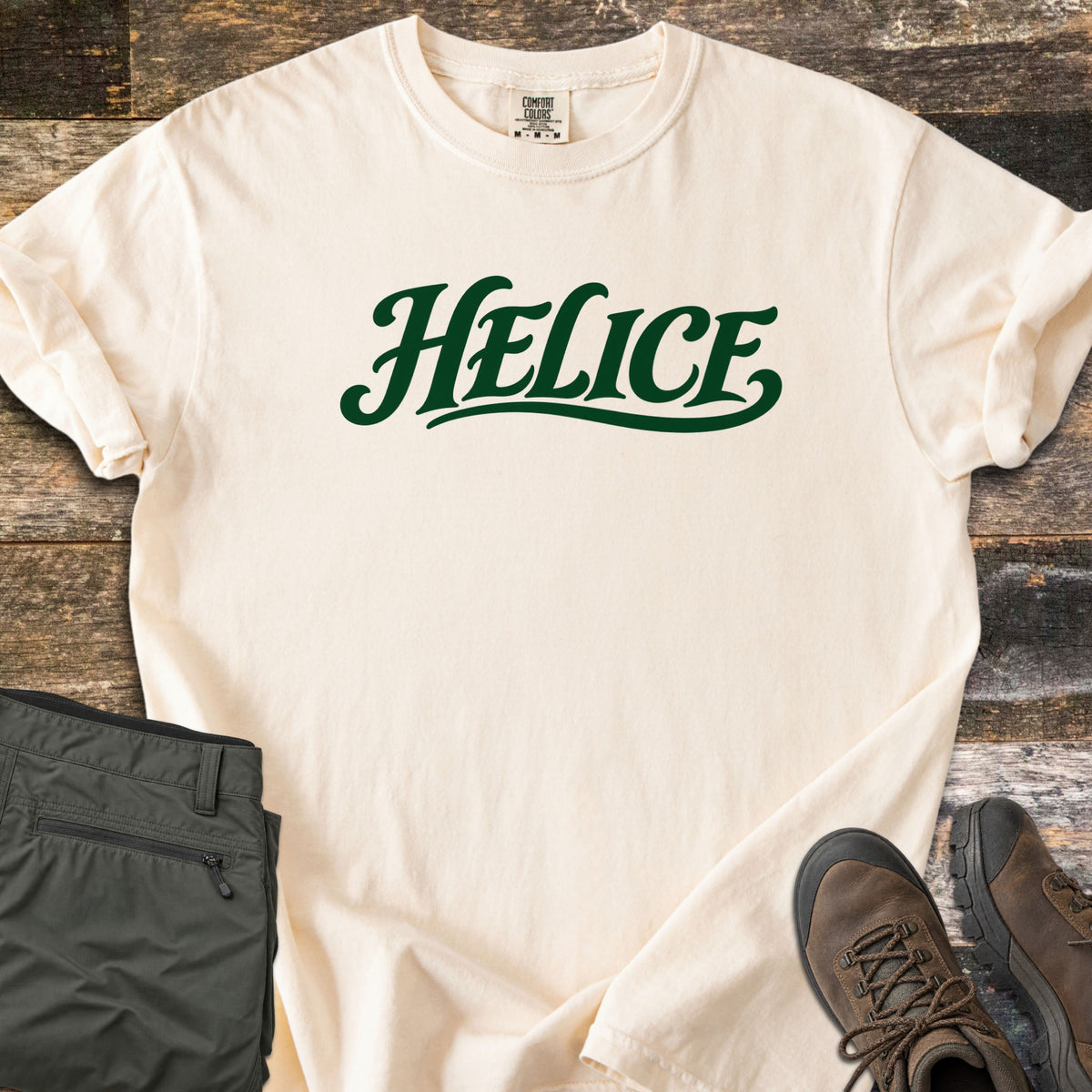 Helice Comfort Colors T-shirt in Ivory, laid flat. The word Helice is printed in dark green script lettering with a swash underline across the chest. Sleeves are rolled, on a rustic wood floor beside folded olive cargo pants and brown leather hiking boots.