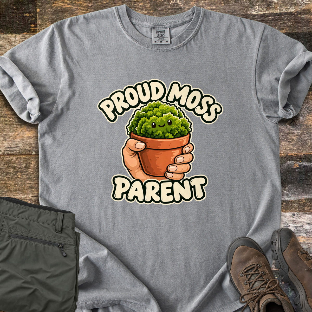 Proud Moss Parent Comfort Colors T-shirt in Grey, laid flat. A cartoon hand holds a terracotta pot of smiling green moss between the words Proud Moss Parent. Sleeves are rolled, with green hiking pants and brown hiking boots on a weathered wood background.