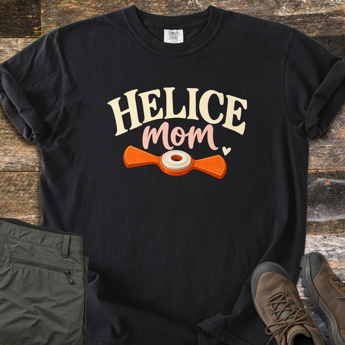 Helice Mom Comfort Colors T-shirt in Black, laid flat. Cream HELICE lettering over pink script mom and a small heart, above an orange helice target with a white center ring. The shirt lies on dark wood planks next to folded olive pants and brown hiking boots.