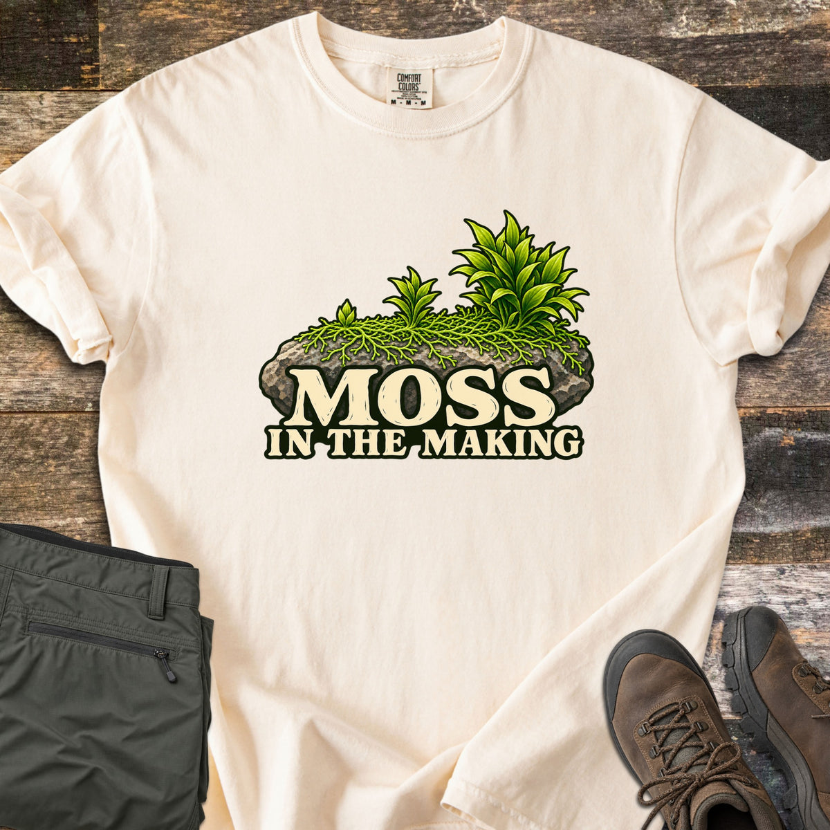 Moss in the Making Comfort Colors T-shirt in Ivory, laid flat. A mossy gray rock sprouting green leafy plants sits above the words Moss in the Making. Olive hiking pants and brown hiking boots lie beside it on weathered wood planks.