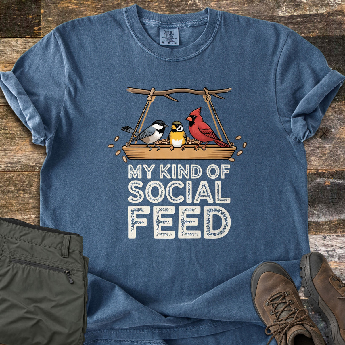 My Kind Of Social Feed Comfort Colors T-shirt in Denim, laid flat. Three cartoon birds, a chickadee, a goldfinch and a cardinal, perch on a hanging seed tray above the words My Kind Of Social Feed. Olive hiking pants and brown hiking boots sit beside it on weathered wood.