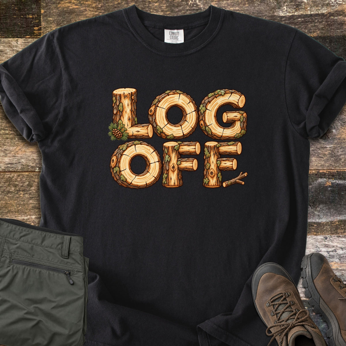 Log Off Comfort Colors T-shirt in Black, laid flat. The words Log Off are spelled in letters made of cut wooden logs with bark, moss and a pine cone. The shirt lies on weathered wood planks beside folded olive pants and brown hiking boots.