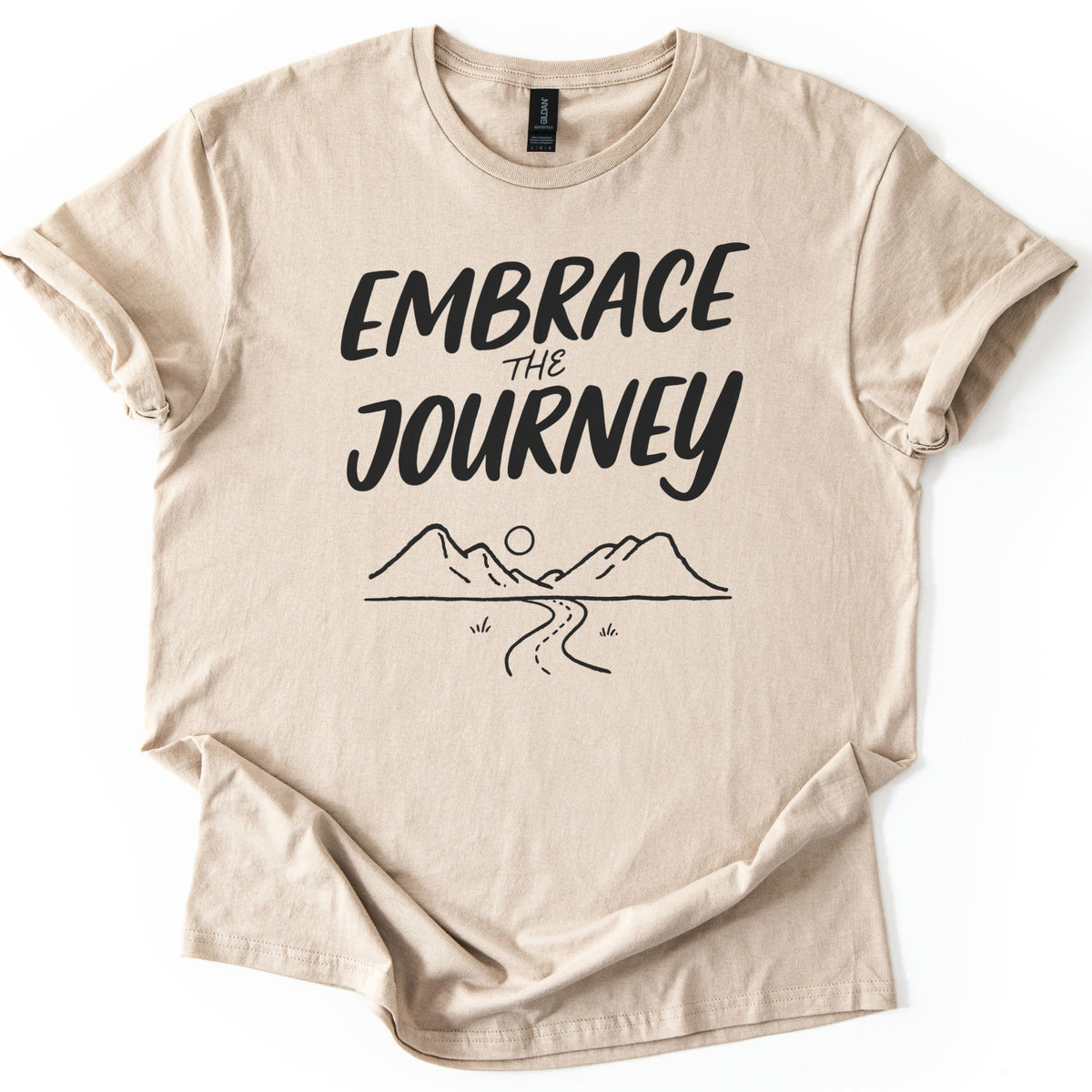 Embrace The Journey Trail Comfort Colors T-shirt in Sand, laid flat. Black hand-lettered words Embrace the Journey sit above a line drawing of two mountains, a round sun, and a winding road with tufts of grass. The sleeves are cuffed, on a white background.
