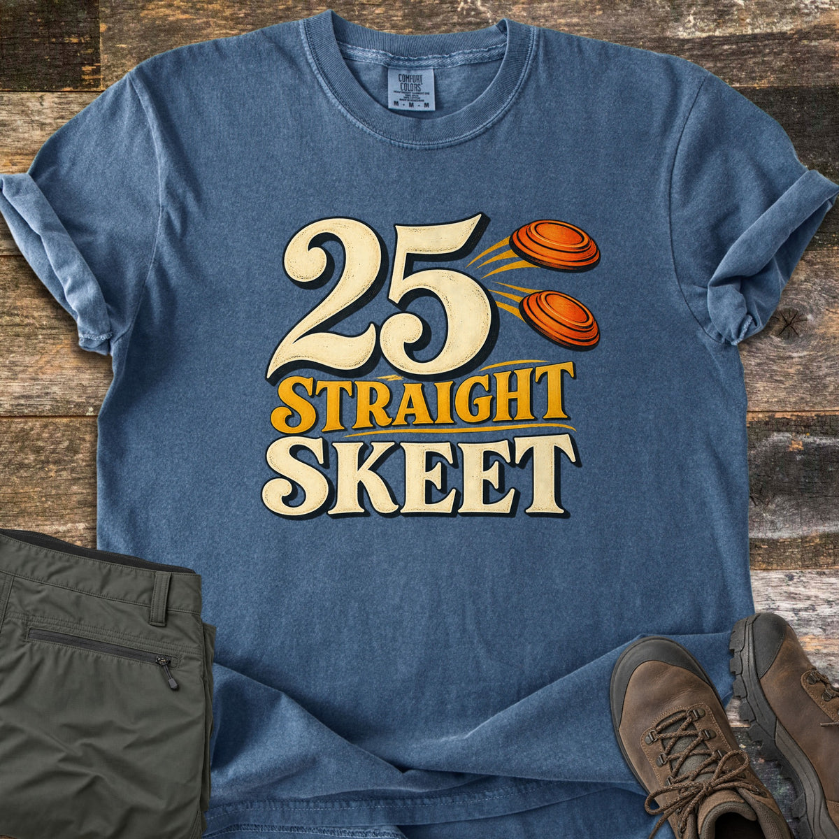25 Straight Skeet Comfort Colors T-shirt in Denim, laid flat. Large cream numbers 25 with two orange clay targets flying beside them, above the words Straight Skeet in gold and cream. Shown on weathered wood with olive cargo pants and brown hiking boots.