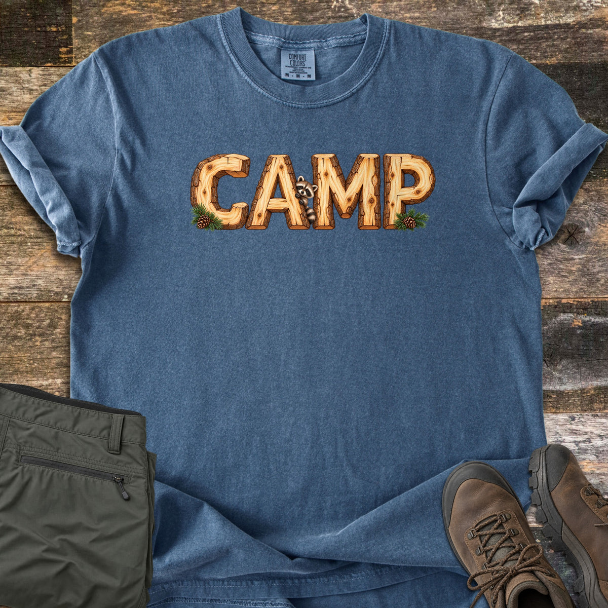 Camp Comfort Colors T-shirt in Denim, laid flat. The word CAMP is spelled in wood log letters with pine sprigs and cones, and a raccoon peeks out beside the A. The shirt lies on weathered wood planks with olive cargo pants and brown hiking boots.