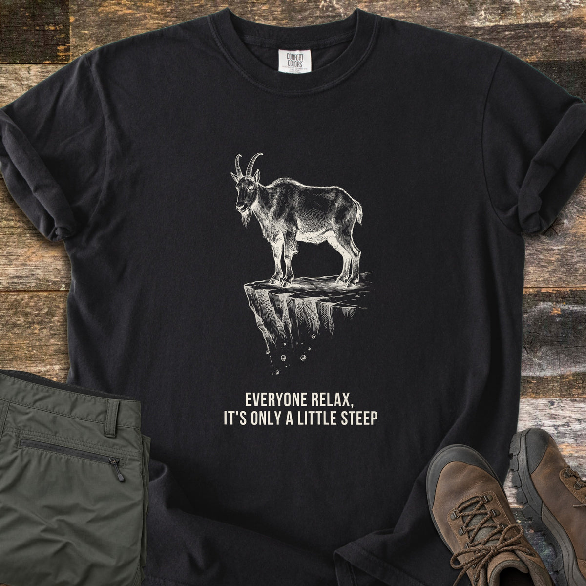 Everyone Relax It's Only A Little Steep Comfort Colors T-shirt in Black, laid flat. A white sketch of a horned mountain goat standing on a crumbling cliff ledge above the words Everyone Relax, It's Only A Little Steep. Sleeves are rolled, on wood planks with green hiking pants and brown boots.