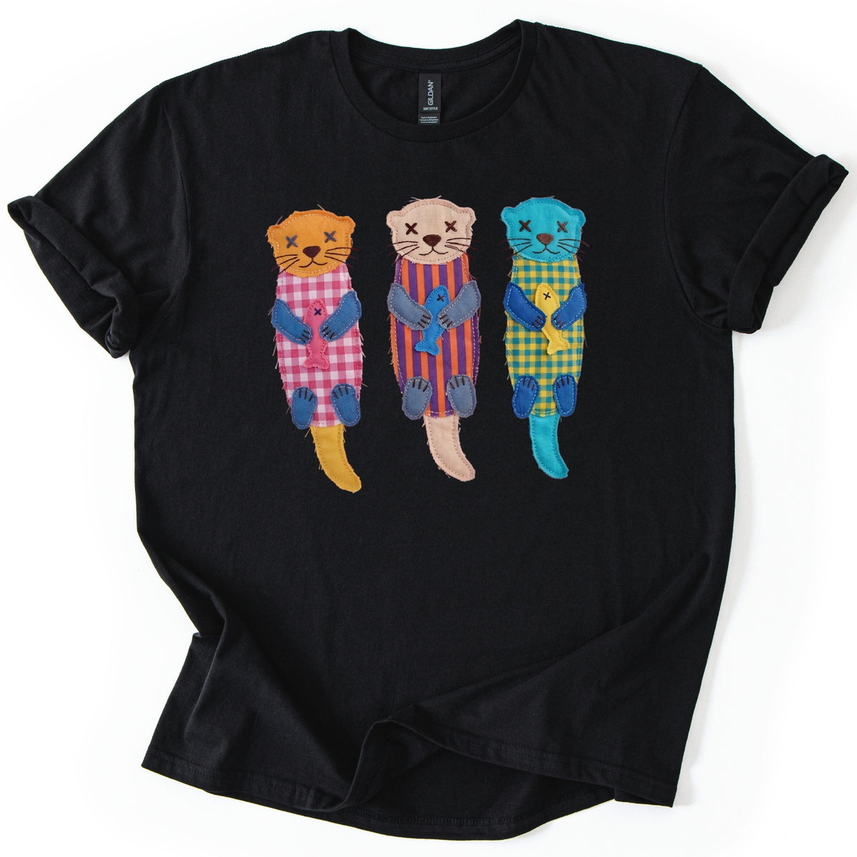 Patchwork Otter Comfort Colors T-shirt in Black, laid flat. Three patchwork otters with X-stitched eyes each clutch a small fish: orange in pink gingham, beige in purple and orange stripes, turquoise in yellow gingham. No words. Rolled sleeves on a white background.